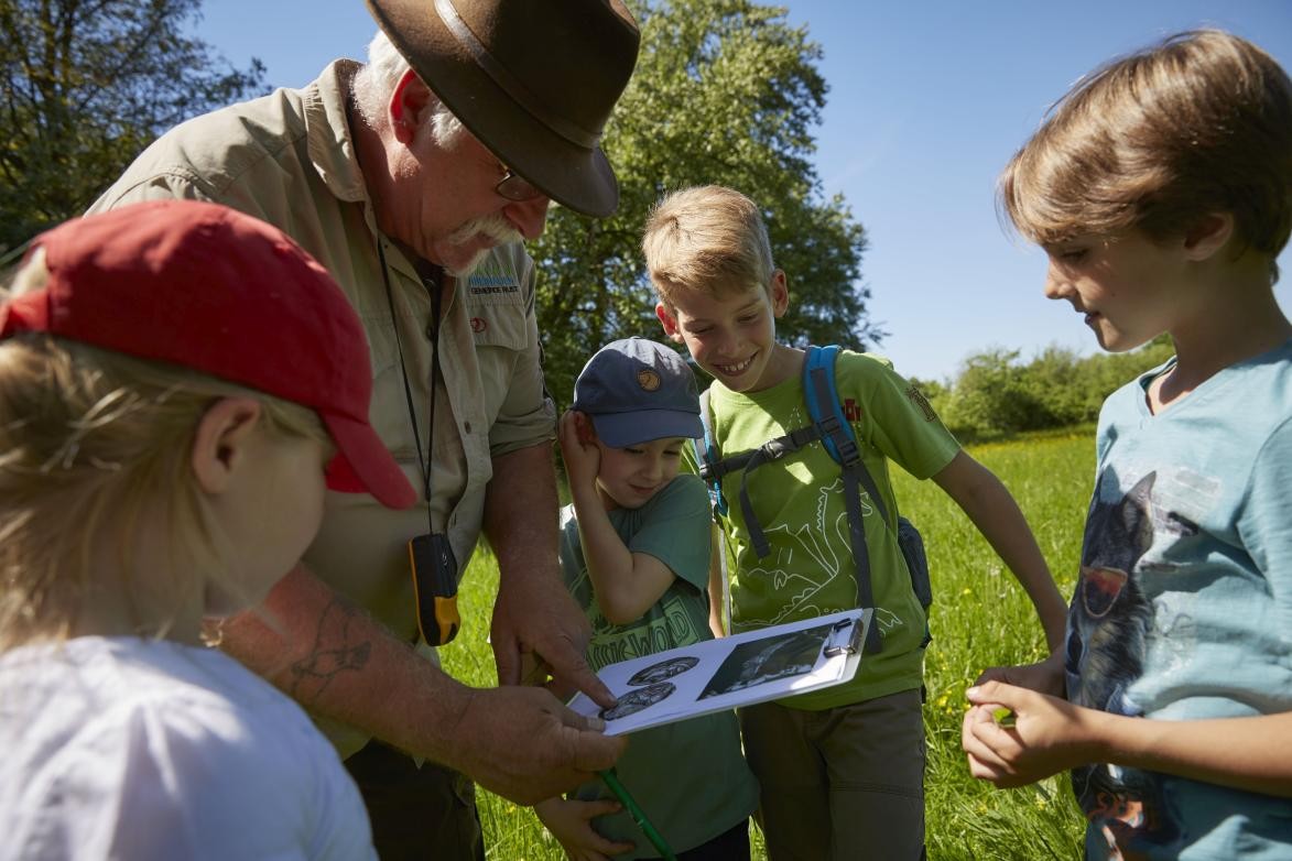 Vier Kinder stehen rund um einen Ranger in der Natur, während er anhand einer Abbildung in seiner Hand etwas erklärt