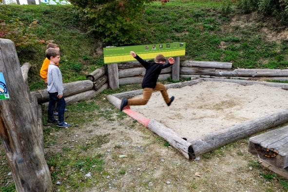 ein Junge springt mit einem Weitsprung in ein Sandbecken, links von ihm stehen zwei weitere Jungs und schauen zu