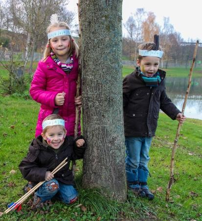 Drei Kinder mit selbstbasteltem Indianerkopfschmuck und Pfeil und Bogen stehen um einen Baumstamm herum