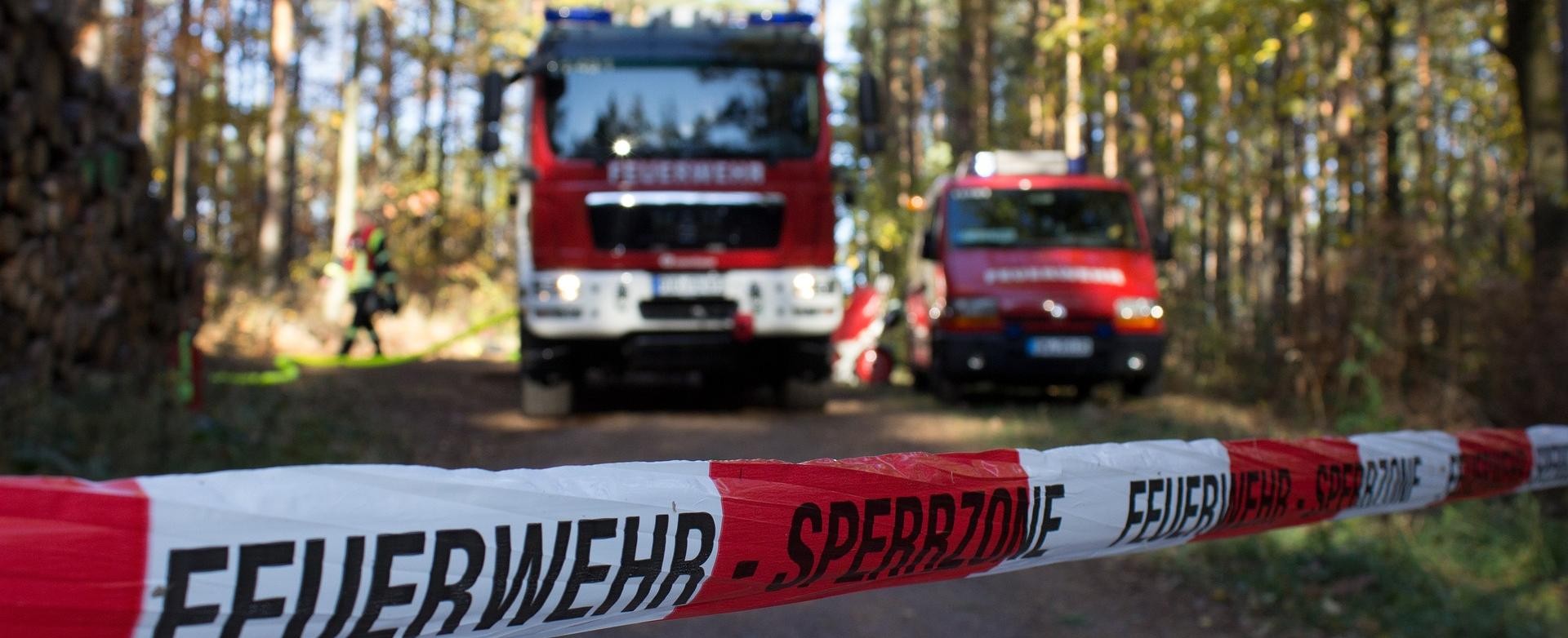 Bevölkerungsschutz Hinter einem Absperrband sieht man zwei Feuerwehrautos auf einem Waldweg