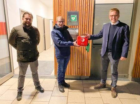 (v.r.) Bürgermeister Kai-Achim Klare, Mitarbeiter Maximilian Gorecky und Amtsleiter Alexander Schindler bei der Anbringung des Defibrillators an der Rheingießenhalle Drei Männer stehen vor dem Eingang einer Halle und halten einen Defibrillator in den Händen