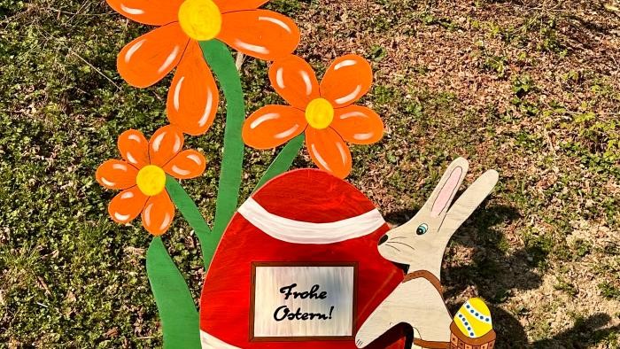 Eine Holzfigur, die aus einem kleinen Osterhasen mit Ei im Korbrucksack, einem großen, roten Ei mit der Aufschrift "Frohe Ostern", einem kleinen, gelben Ei und mehreren Blumen besteht